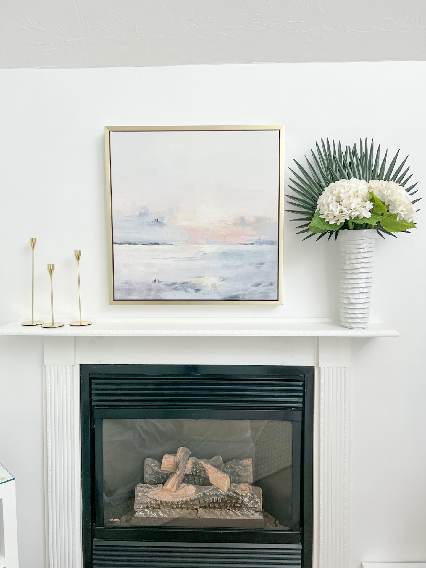 fireplace with abstract sunset artwork hung over it, and a vase with palm stems & hydrangeas